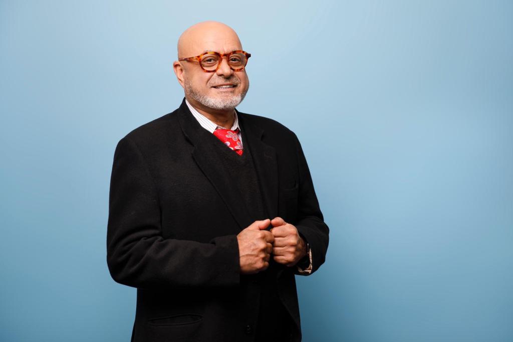Portrait of Sassan Zabeti — Founder, Zabeti Life Academy Portrait of Sassan Zabeti standing against a light blue background, wearing a black suit, white shirt, red patterned tie, and round glasses, with a confident, warm smile.
